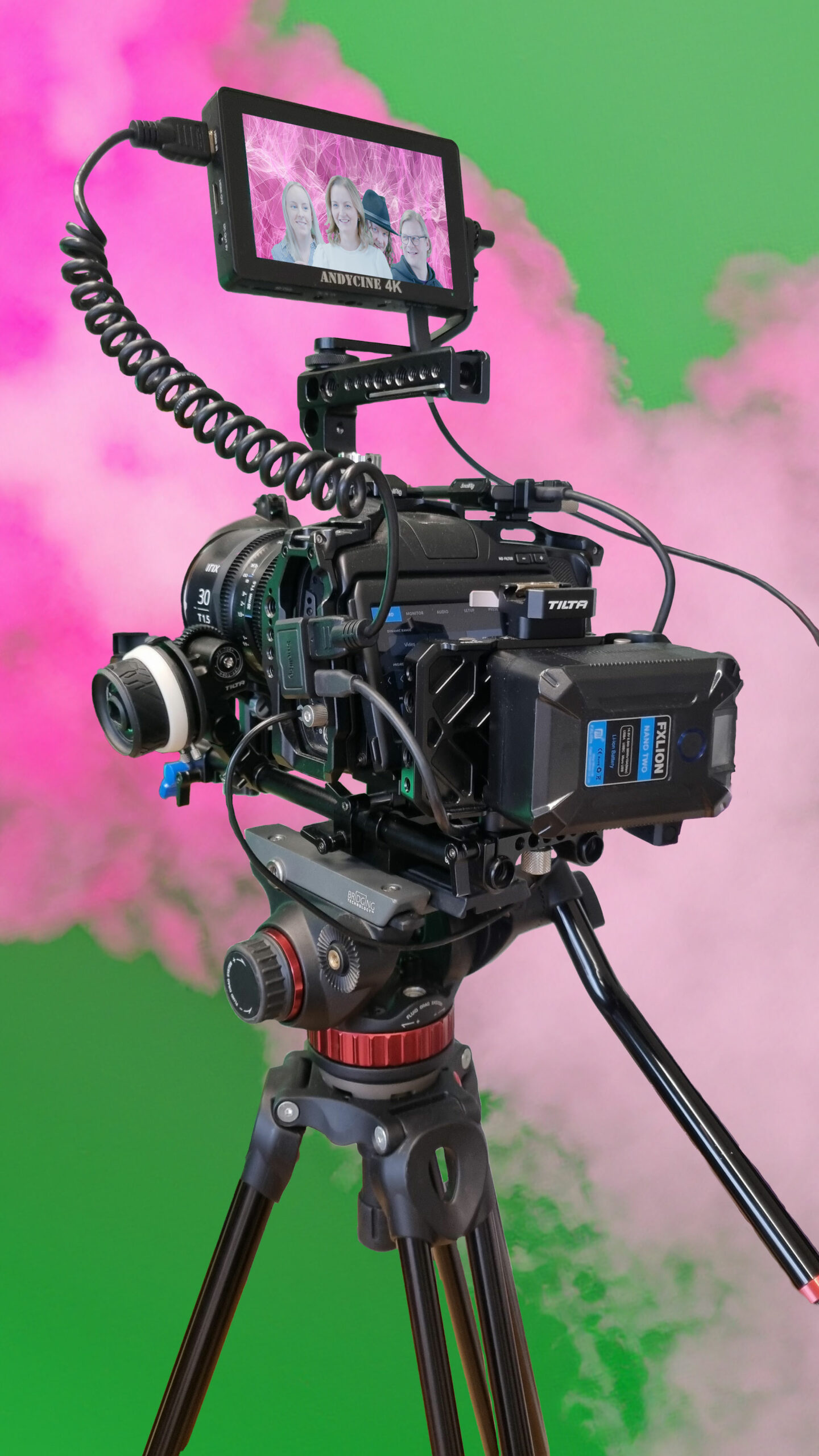 Videokamera jalustalla. näytöllä ryhmä ihmisiä. Taustalla nousee pinkkiä savua.